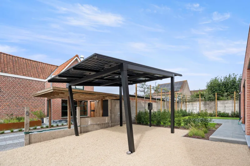 Zwarte stalen solar carport met negen zonnepanelen en een Etrel-laadpaal op sokkel bij een woning in Torhout
